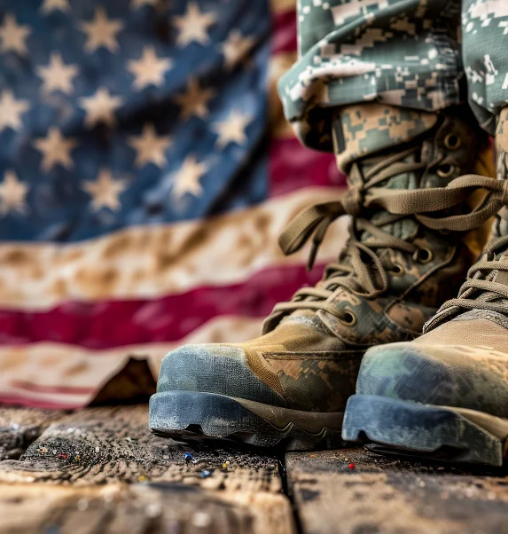 Close-up of military boots on a rustic surface, with a blurred American flag in the background